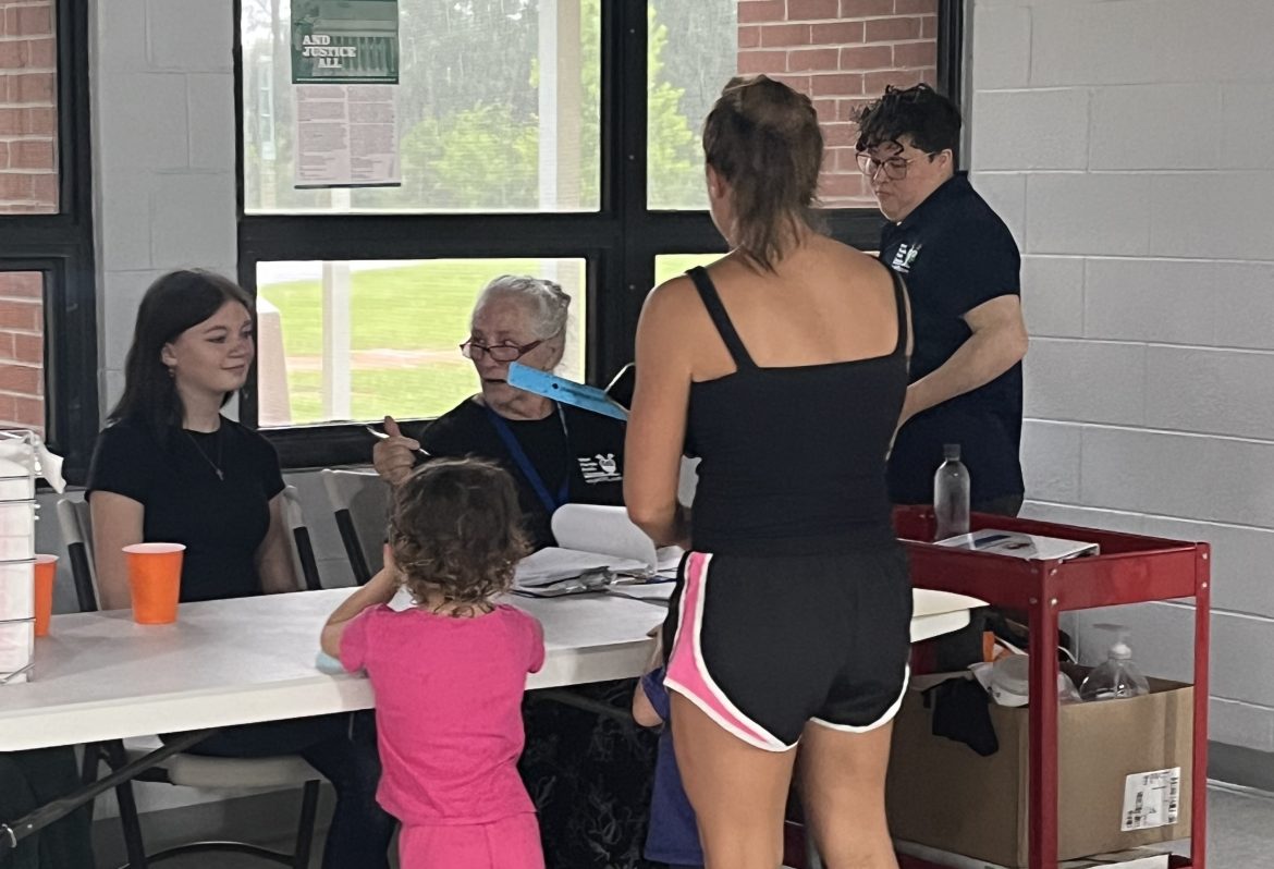 Pais e filhos esperam na fila para serem atendidos em uma mesa dobrável em um local de refeições de verão. 